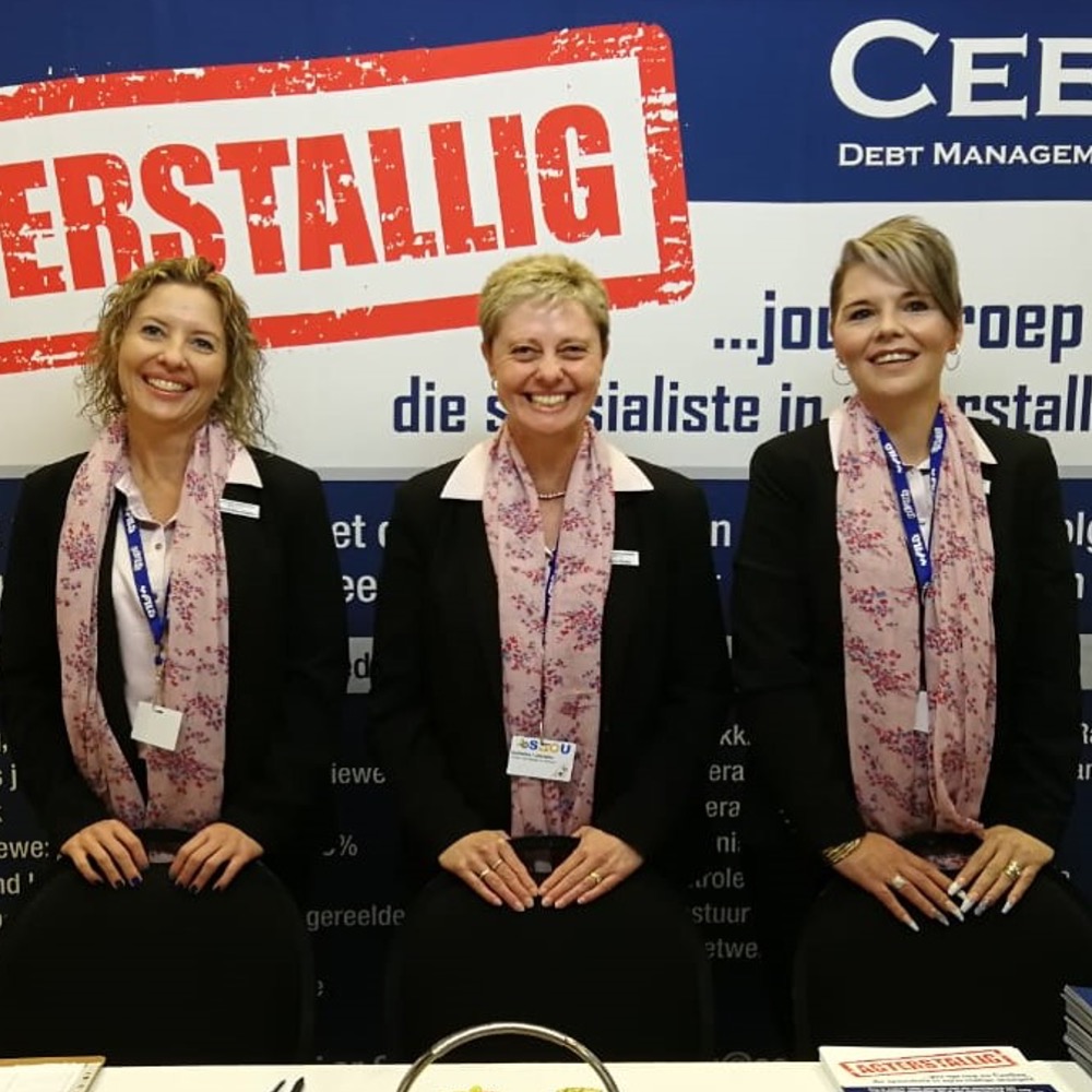 Three smiling women in black blazers with pink floral scarves sit behind a conference booth, badges visible, a blue banner with a red 'ERSTÄLLIG' stamp behind them
