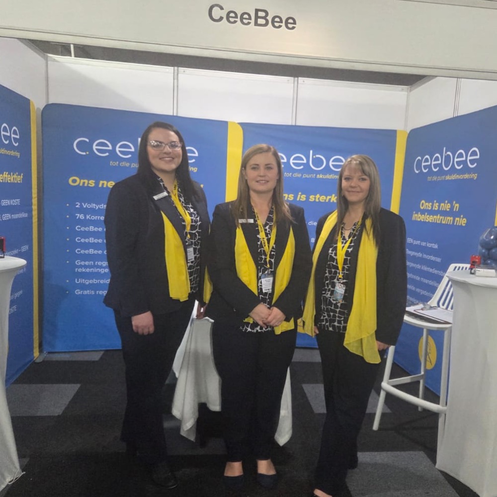 Three women in black outfits with yellow scarves pose at a CeeBee trade show booth with blue banners behind them.