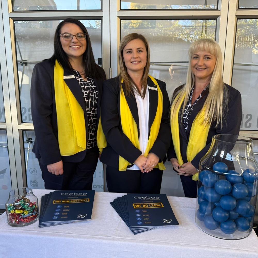 Three smiling women in navy outfits with yellow scarves stand behind a display table with blue brochures and jars of blue balls.
