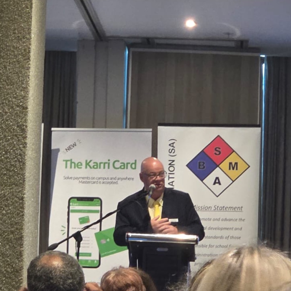 Bald man in a dark suit and yellow tie speaking at a podium, with banners for The Karri Card and a diamond-shaped logo behind him.