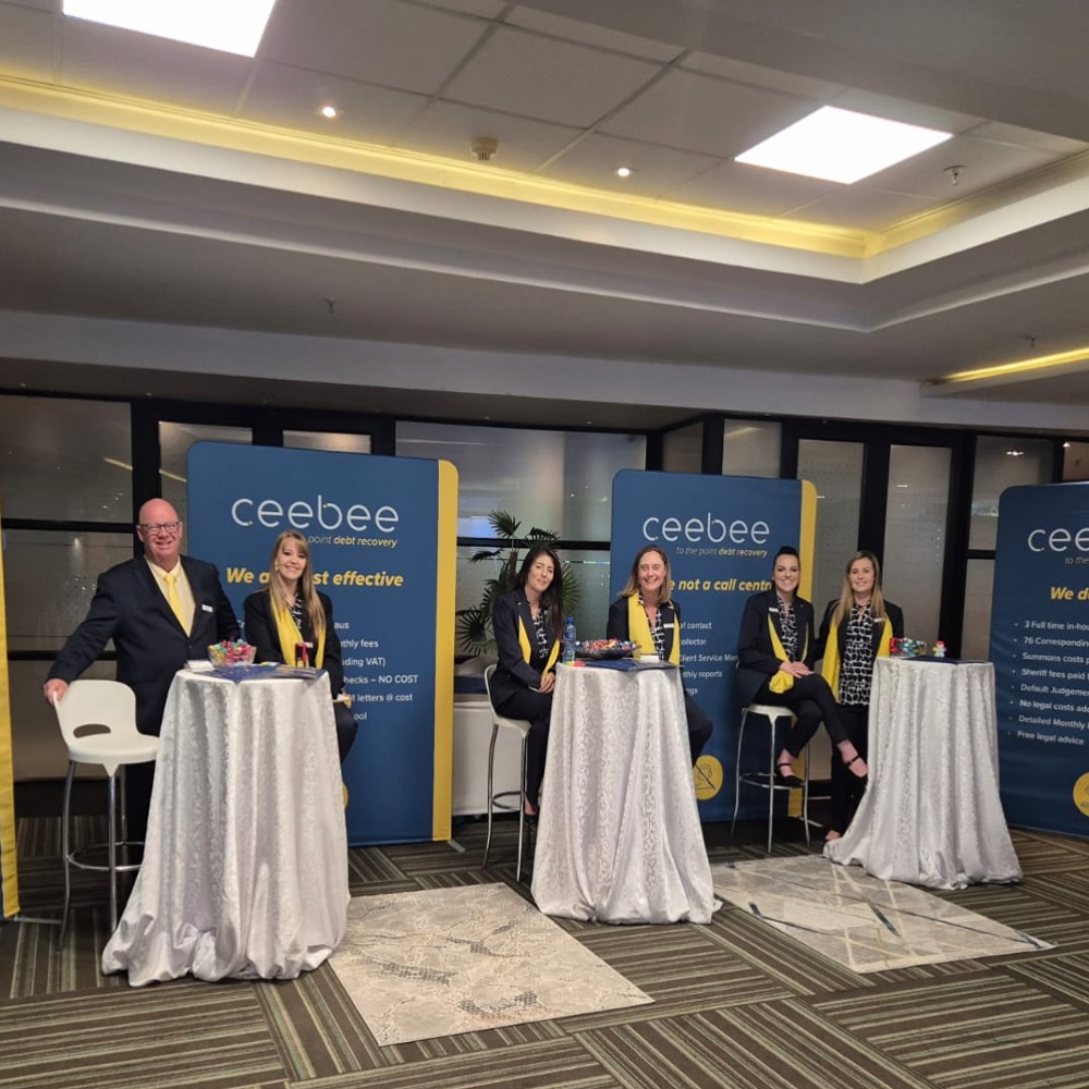 Group of five professionals standing and sitting behind round, high-top tables with silver tablecloths at a conference booth for Ceebee debt recovery, wearing yellow scarves and business attire.