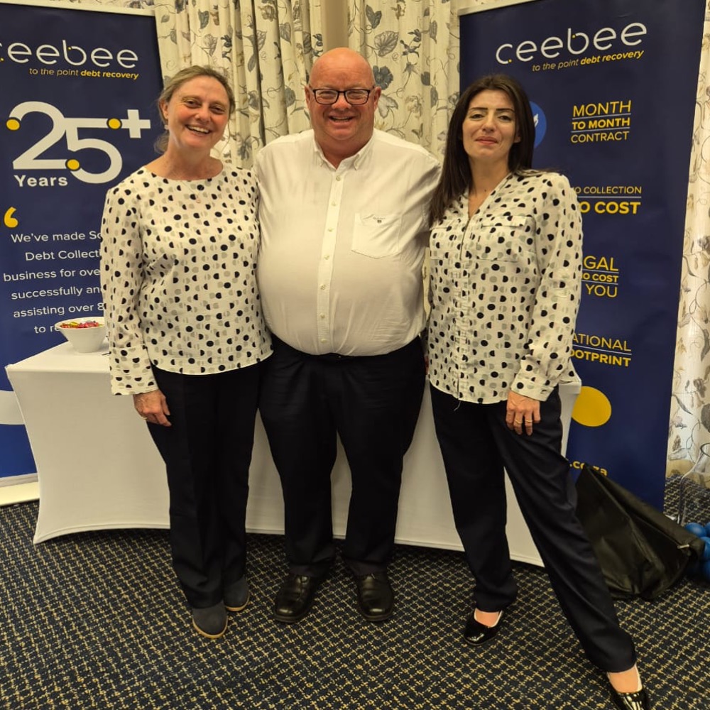 Smiling trio posing in front of blue debt-recovery banners at an event. Two women on either side of a man in a white shirt and black pants.