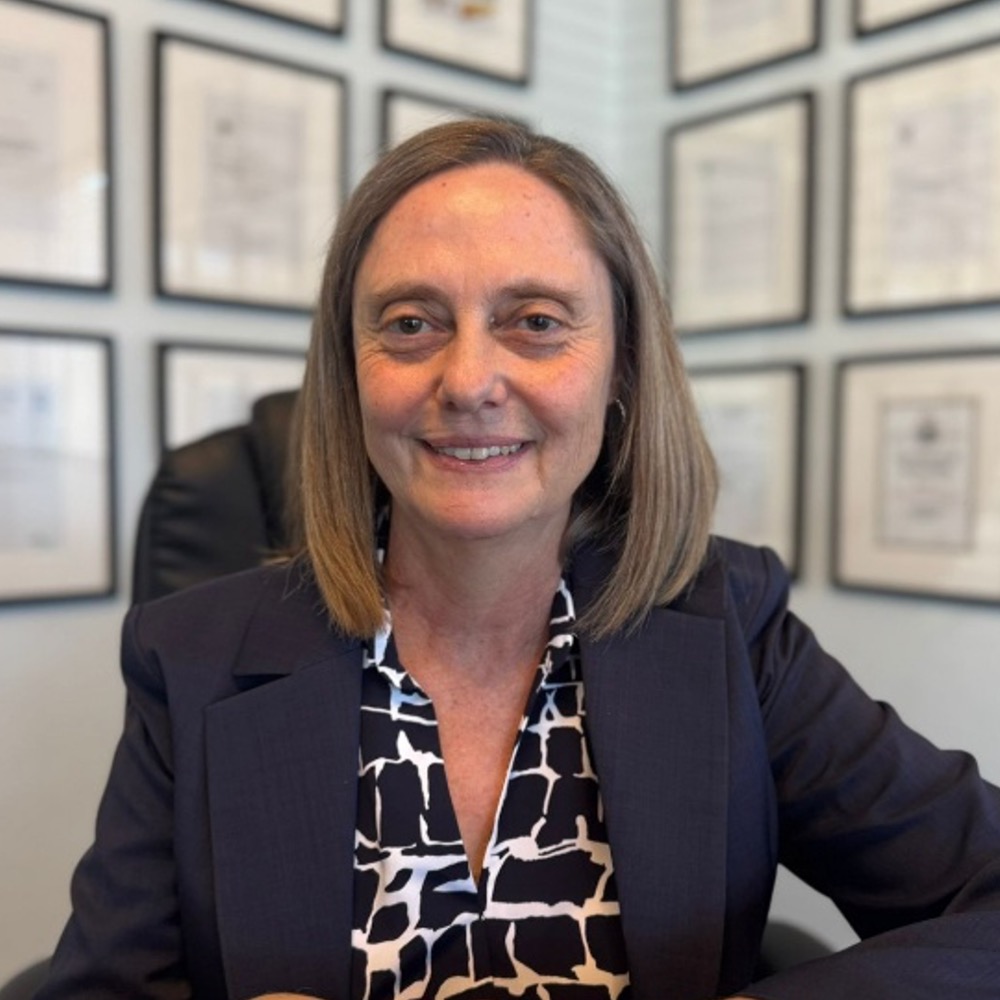 Smiling woman with shoulder-length gray hair, wearing a dark blazer and patterned blouse, in an office with certificates on the wall.