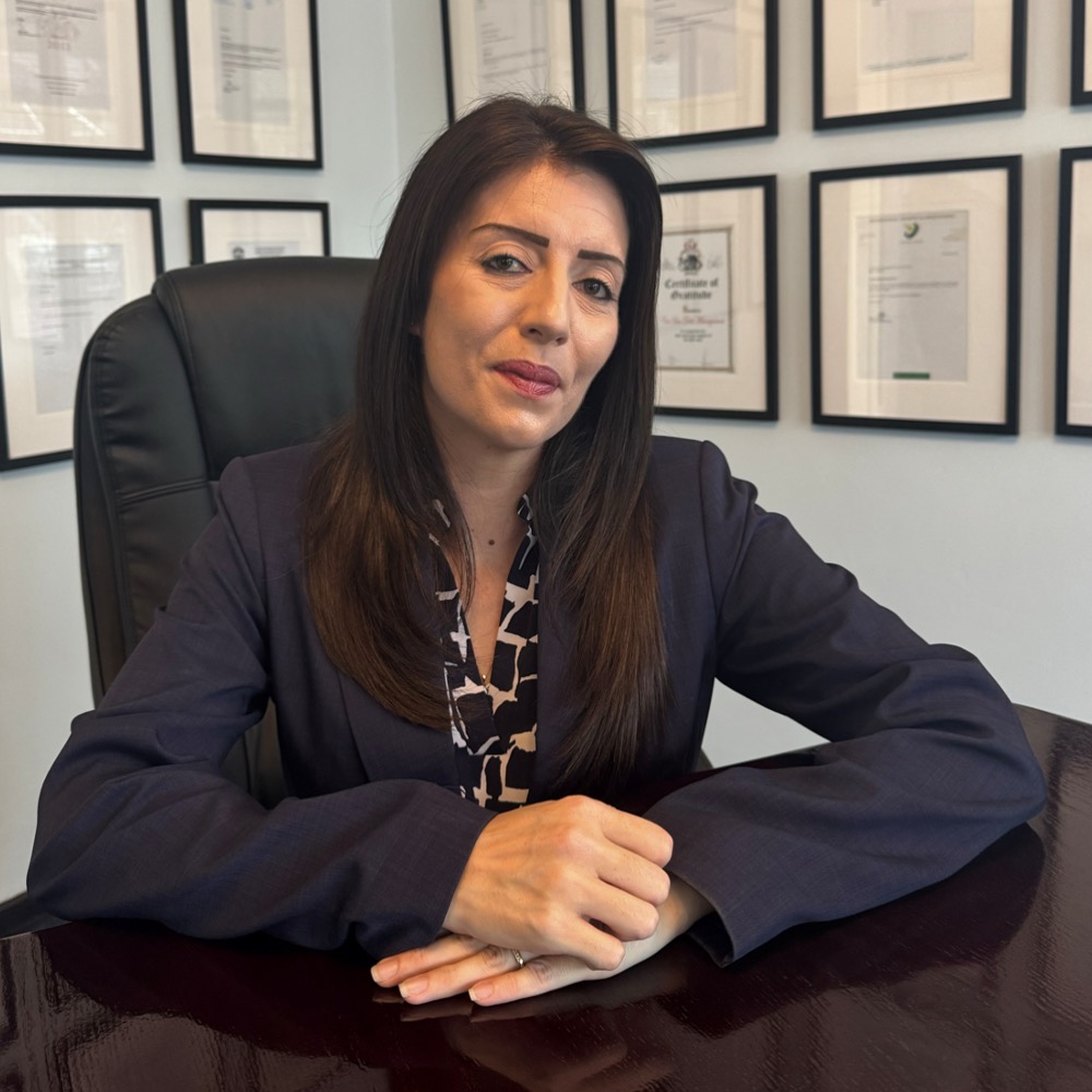 Professional woman seated at a dark wood desk in a navy blazer, hands folded, with framed certificates on the wall behind her.