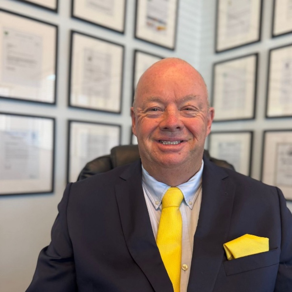 Smiling bald man in a navy suit with a yellow tie and pocket square, in an office with framed certificates on the wall.