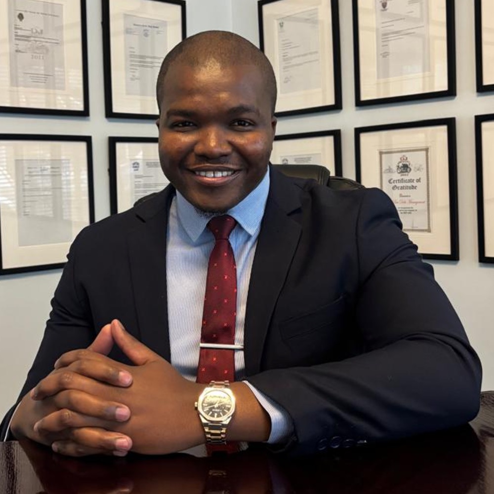 Smiling man in a dark suit and red tie sits at a desk with framed certificates on the wall behind him, conveying a professional office setting.