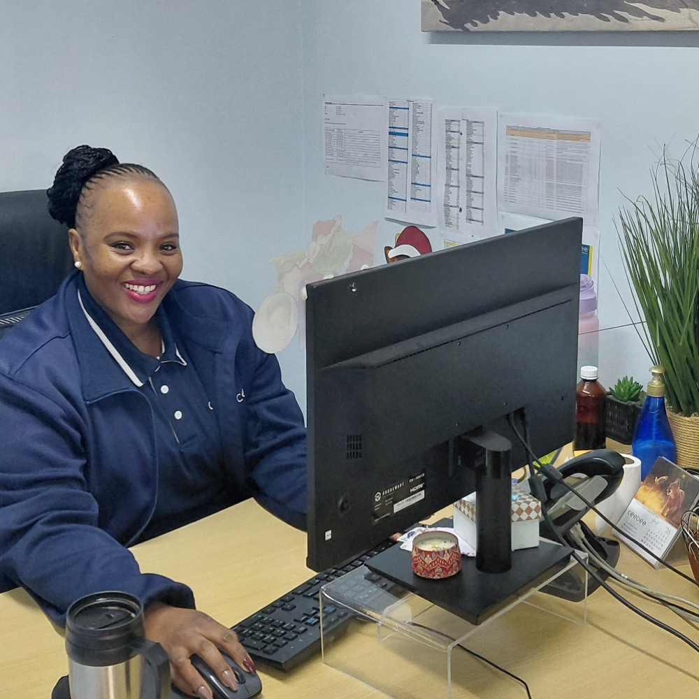 Smiling woman in a blue jacket sits at a desk with a computer monitor and keyboard in an office setting.
