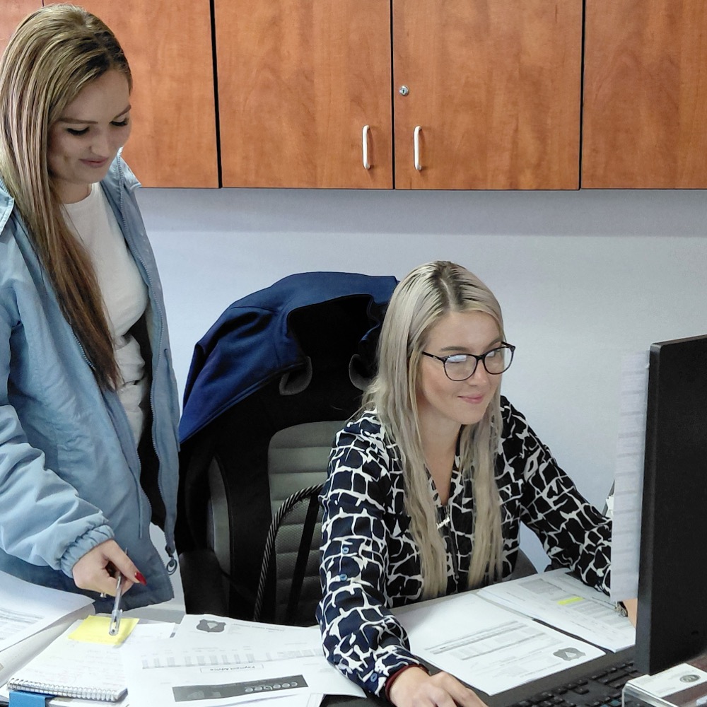 Two women collaborate at a cluttered desk: one standing with a pen over documents, the other seated at a computer looking at papers.