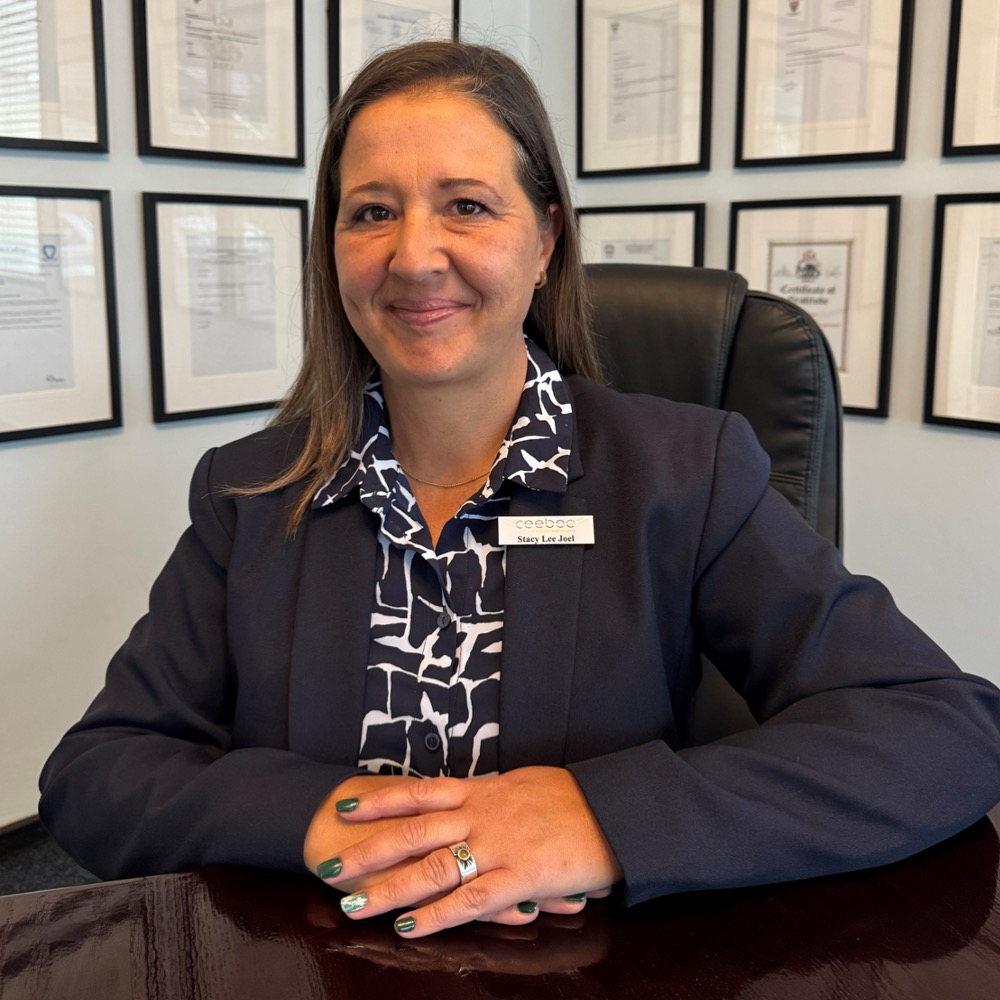 Smiling professional woman seated at a desk in an office, wearing a dark blazer and patterned blouse with a visible name badge.