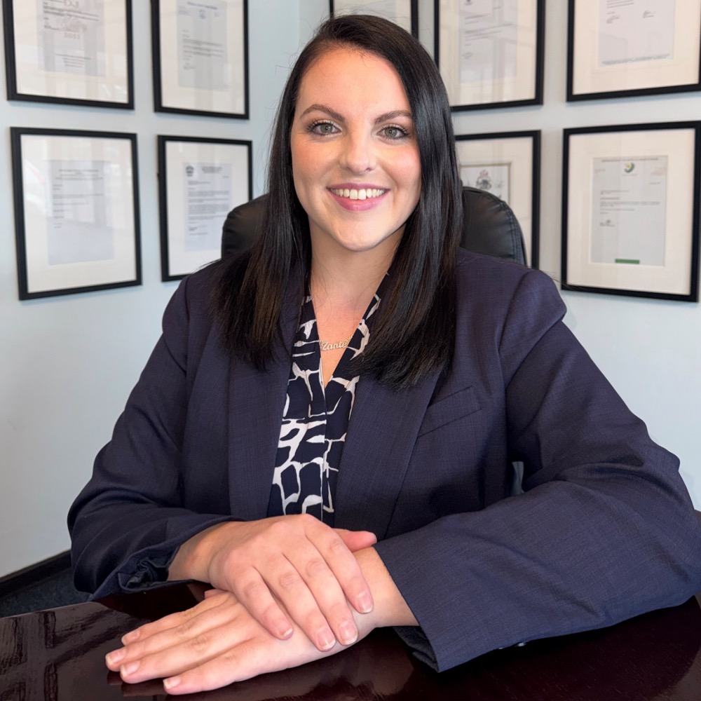 Smiling woman with dark hair in a navy blazer seated at a desk in a professional office, framed certificates on the wall behind.