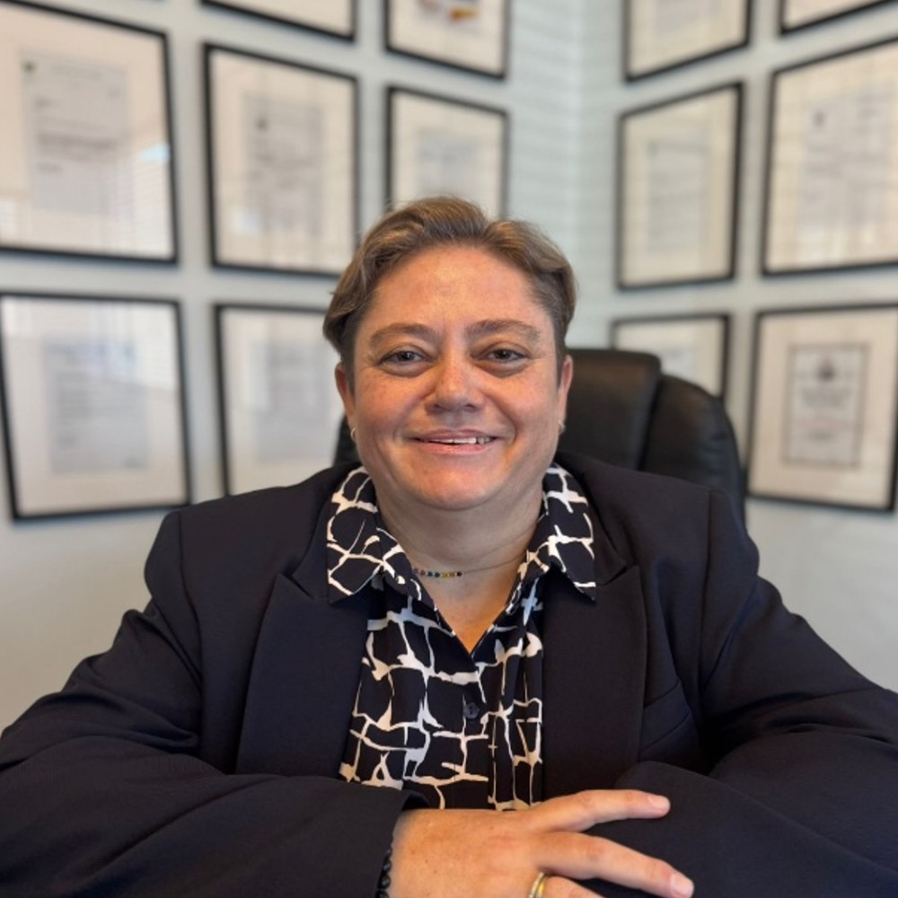 Professional woman in a blazer sits at a desk, smiling, with framed certificates on the wall behind her in an office setting.
