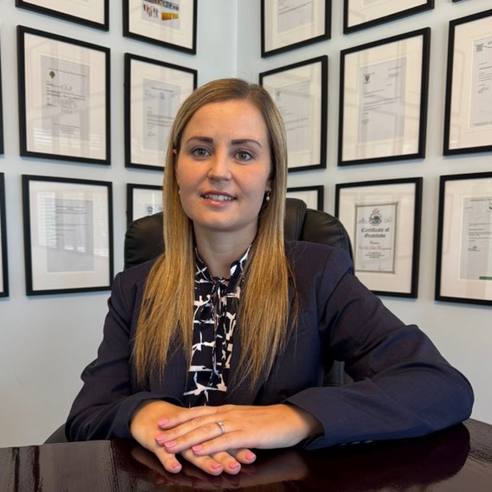 Professional woman in a navy blazer sits at a desk, framed certificates on the wall behind her.