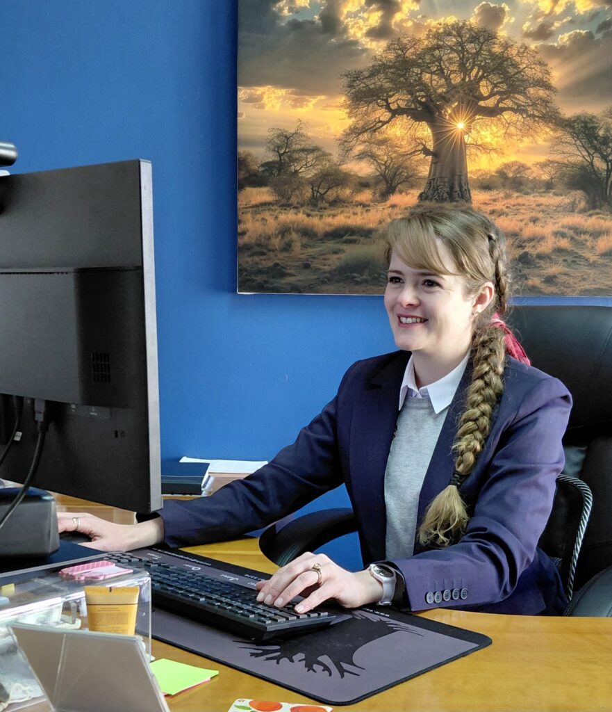 Smiling woman with braided hair typing at a desk; blue wall and a sunset savanna tree poster behind her.