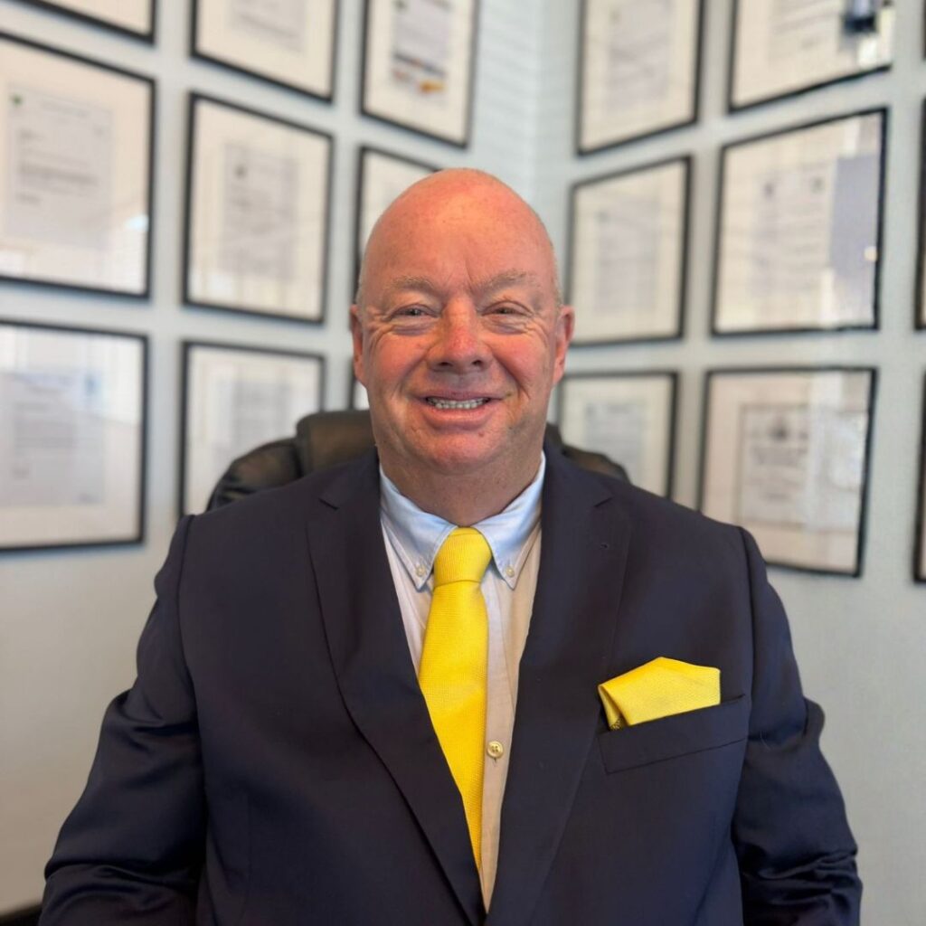 Smiling businessman in a navy suit with a bright yellow tie sits in an office with framed certificates on the wall behind him.