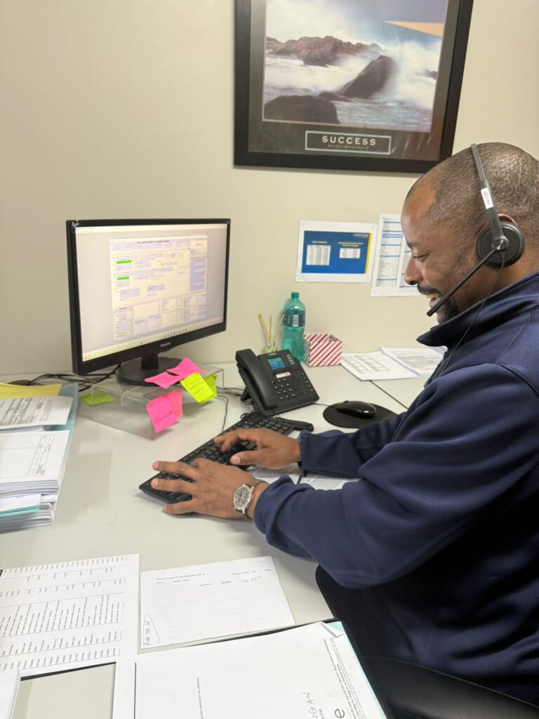 Man with headset typing on a keyboard at a desk in a small office, surrounded by papers, sticky notes, and a monitor.
